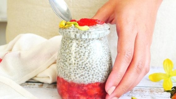 chia seed pudding with strawberry puree, hand holding a spoon, apron, kitchen towel, flowers, pistachios and fresh strawberries