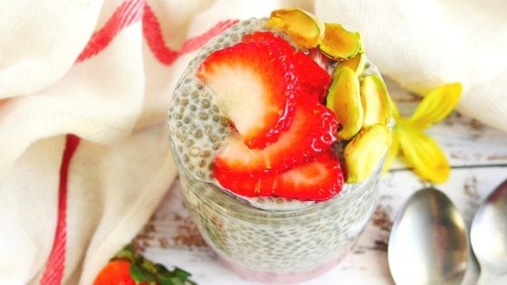 close-up of chia seed pudding with strawberry puree, fresh strawberries, pistachios, two spoons, yellow flowers, kitchen towel and enamel tray