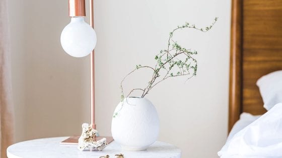 faux flowers in a white flower vase on top of a night stand. Lamp on the side. Faux foliage