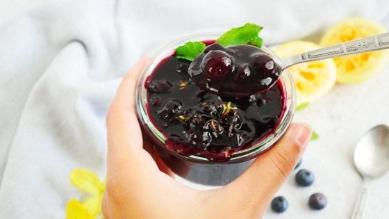 blueberry lemon sauce served in a glass jar with spoon in hand, fresh blueberries, lemons and a second spoon on the table near a kitchen towel