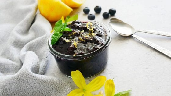blueberry lemon sauce in a glass jar with spoons on the side, fresh blueberries, lemons, kitchen towel and yellow flowers