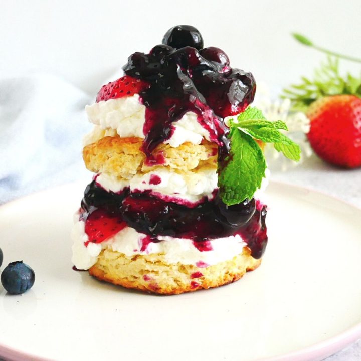 strawberry shortcake with lemon biscuits, blueberry lemon sauce, fresh strawberries in a white and pink plate, fresh blueberries, utensils, and a kitchen napkin